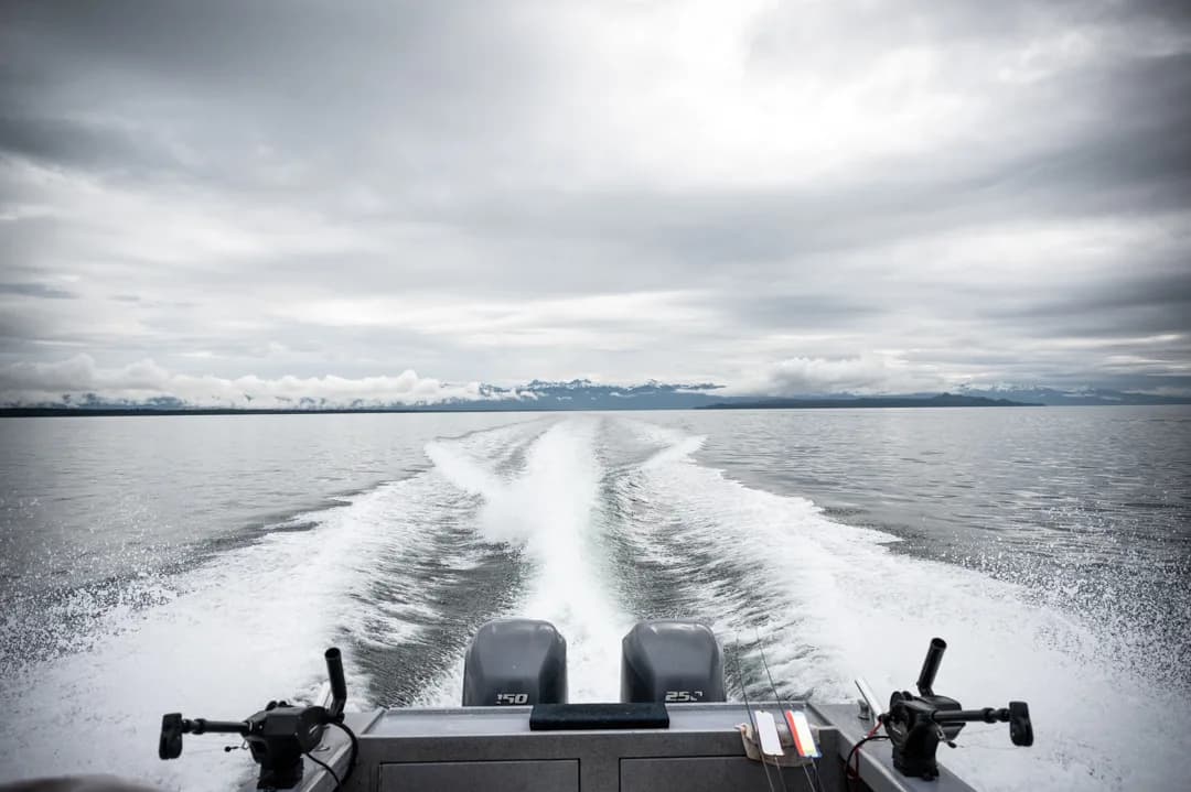 Twin outboards trailing a white wake across Icy Strait, snow-capped mountains on the horizon under overcast skies.