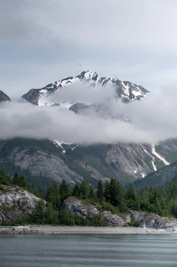 Snow-capped peak rising above low clouds over a rocky shoreline and spruce treeline, Glacier Bay area