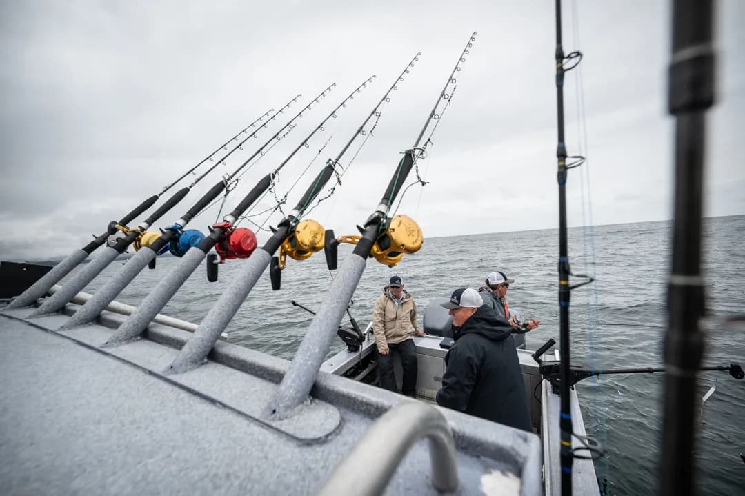 Row of heavy deep-sea rods racked on boat gunwale, three anglers on deck under overcast sky, open water behind.