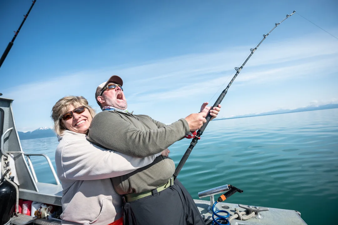 Man and woman on deck, man straining on bent rod with mouth open, calm green water and snow-capped mountains behind