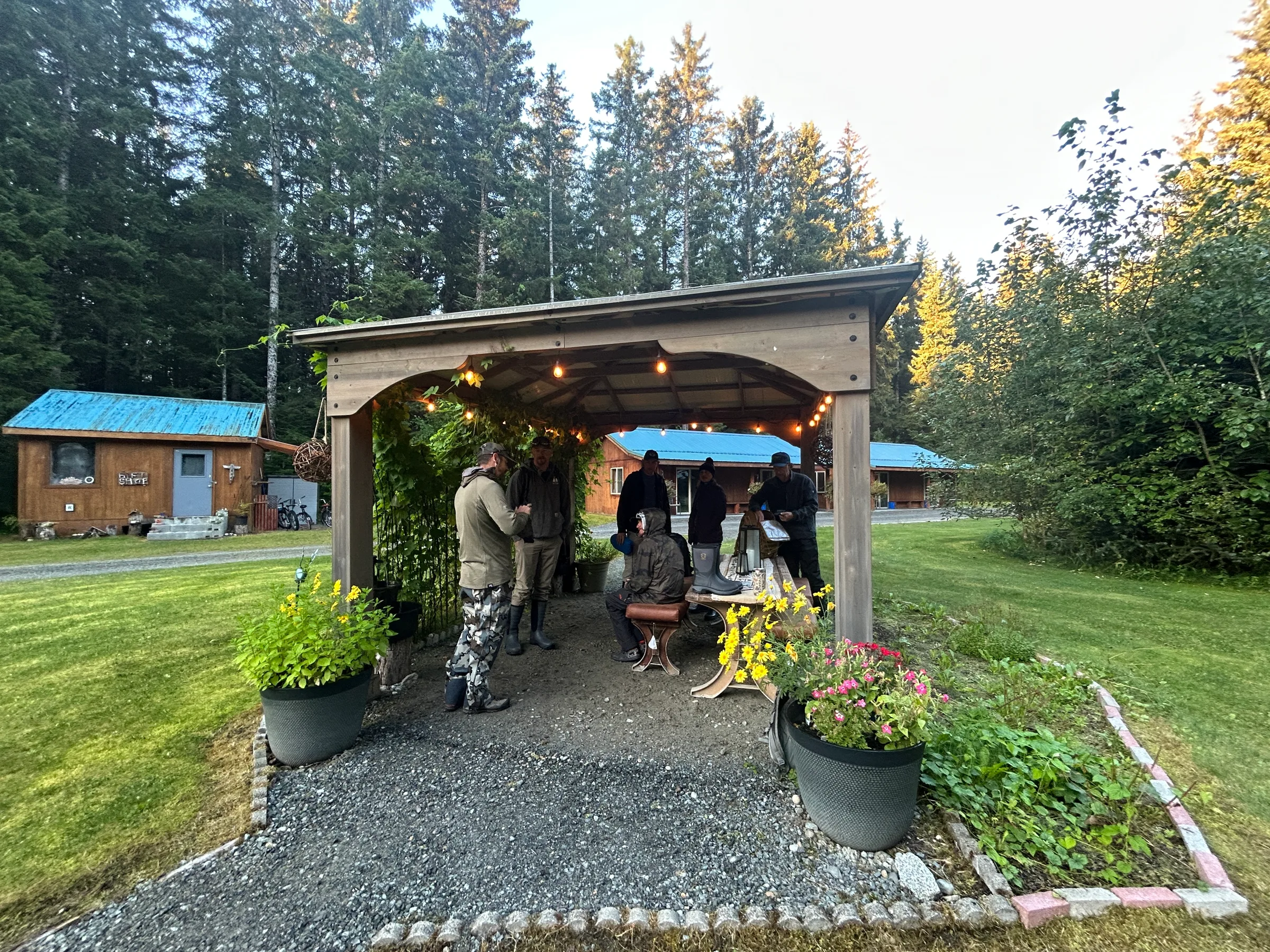 Anglers in waders gather under a wood-framed covered pavilion with string lights, lodge cabins and spruce forest in background.