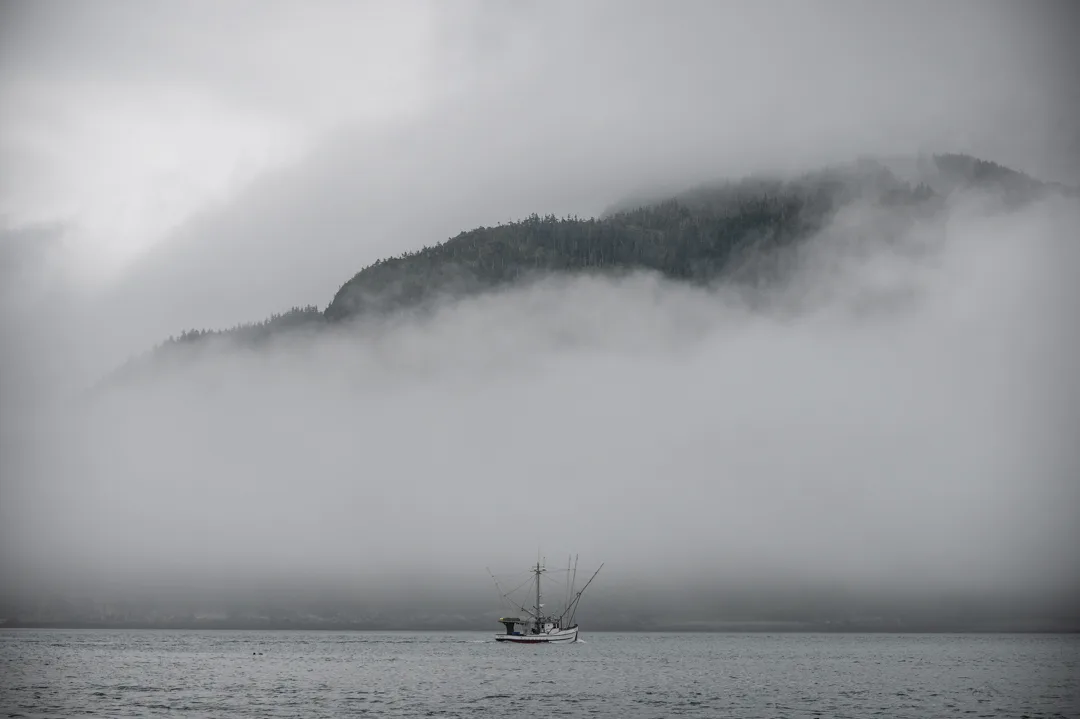 Small fishing vessel crossing fog-wrapped water, forested mountain ridge emerging through low clouds above.