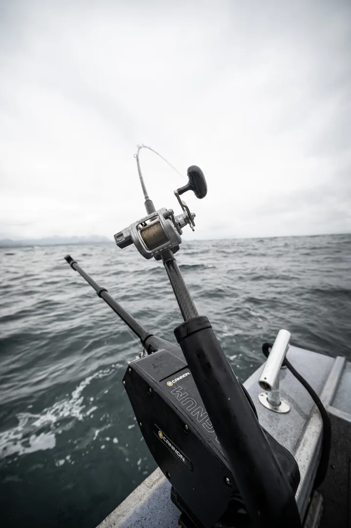 Cannon downrigger and rod with conventional reel mounted on aluminum boat rail, open water and overcast sky behind.