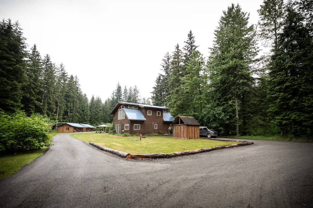 Main lodge building and outbuildings set in a spruce forest clearing, viewed from the gravel driveway.