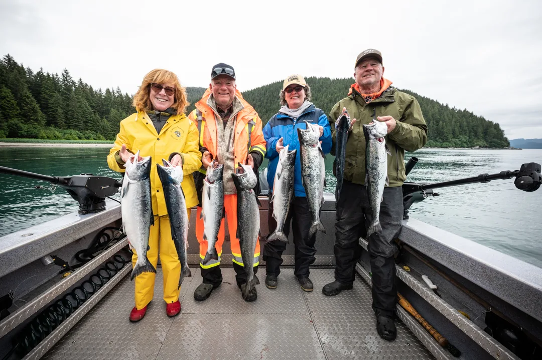 Four anglers on boat deck holding silver salmon, forested Alaskan coastline and calm water in background.