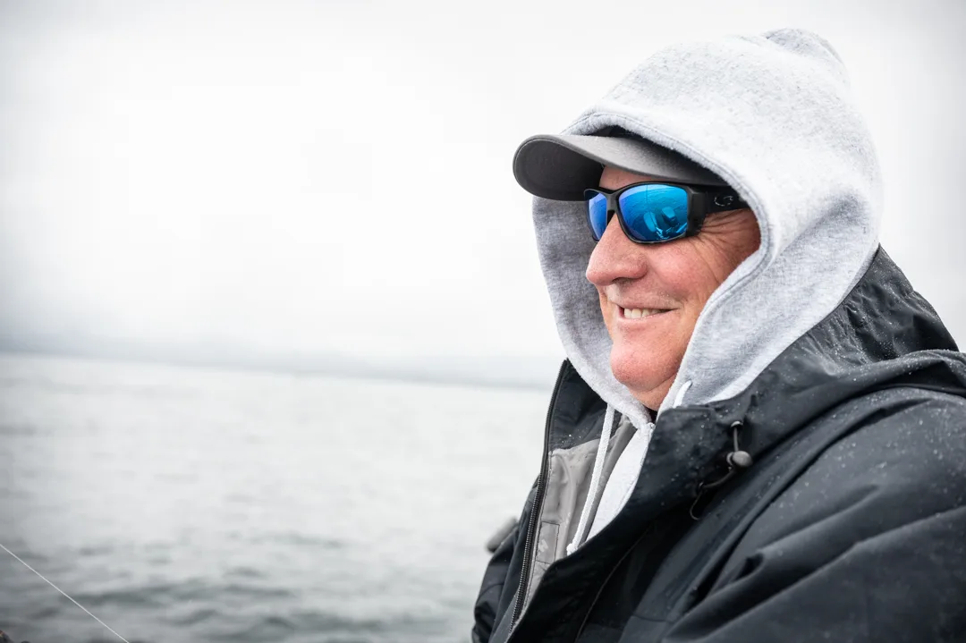 Angler in rain gear and polarized sunglasses smiling on the water, overcast skies behind him.