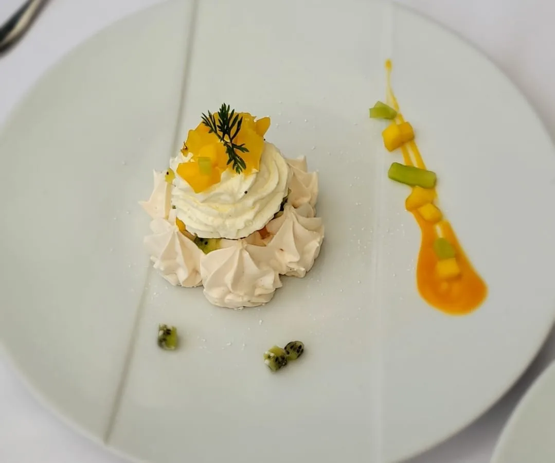 Pavlova with whipped cream, mango coulis, kiwi, and fresh herb garnish plated on white ceramic
