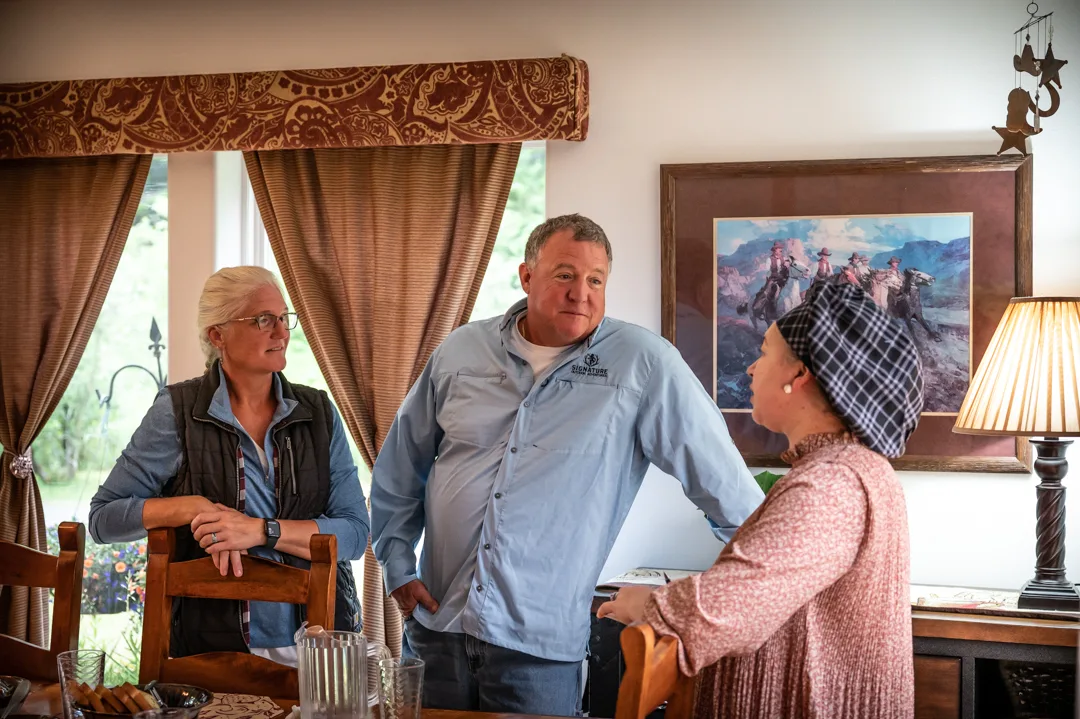 Three guests conversing in a lodge dining room, warm lamp and framed Western art visible in background.