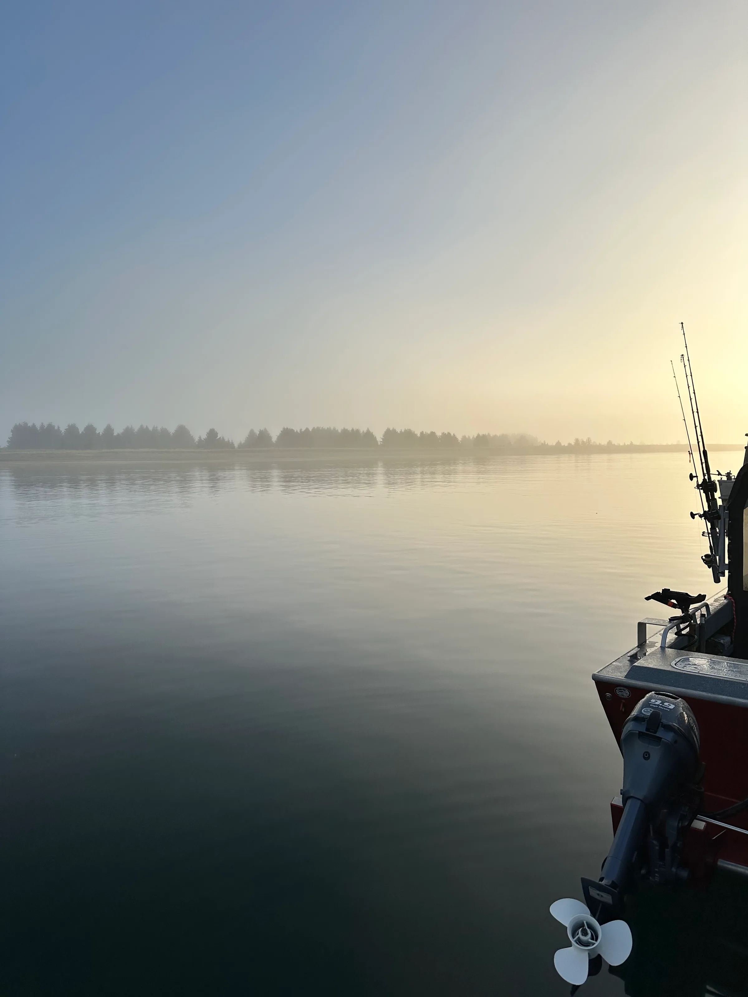 Fishing boat bow and rods at rest on glassy water, forested shoreline dissolving into morning fog, warm sunrise light.