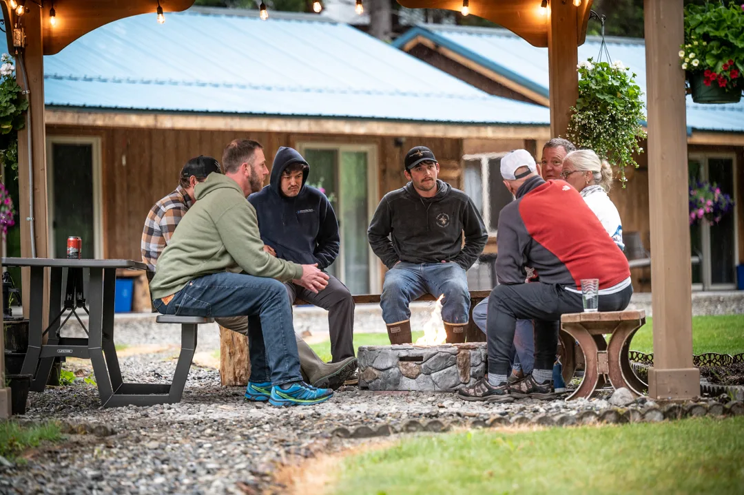 Group of guests gathered around an outdoor stone fire pit under a pergola with string lights, lodge building behind