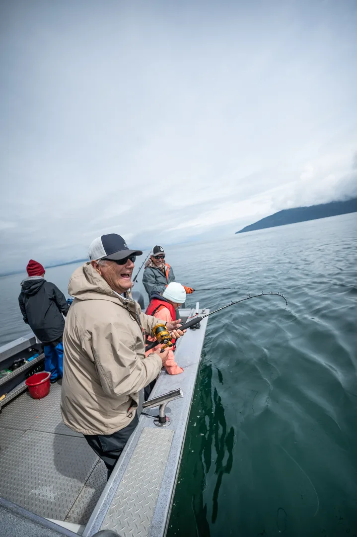 Angler working a bent rod off the stern rail, three others fishing alongside on calm Alaskan water, coastal ridge in the distance.