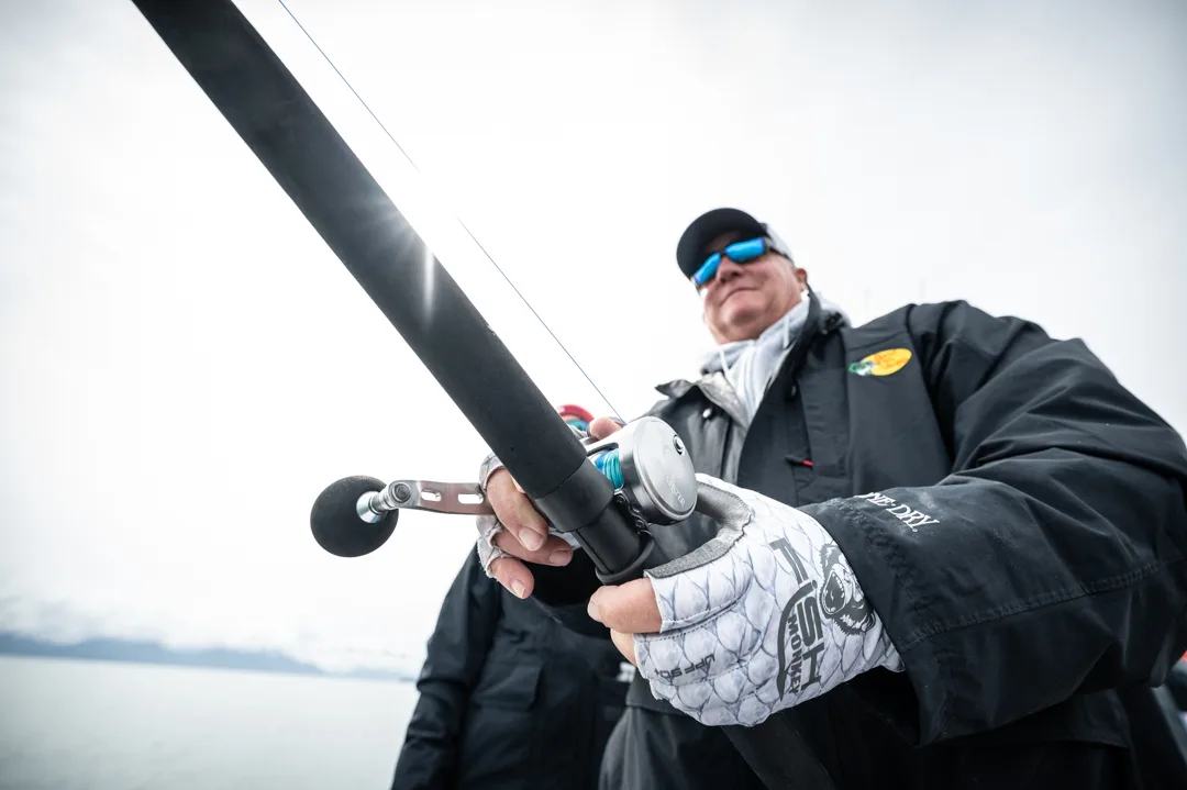 Angler in fingerless gloves working a heavy reel on a saltwater rod, overcast Alaskan water visible behind.