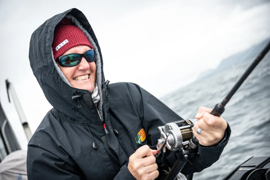 Woman in rain gear and red beanie working a rod and reel on a grey overcast day on the water.