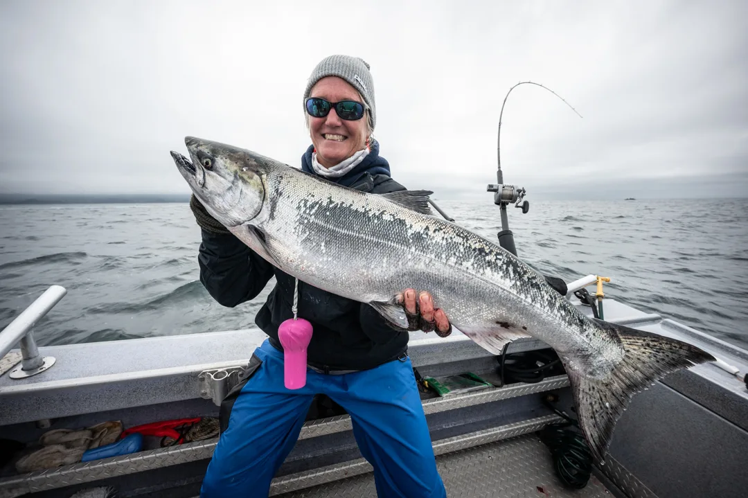 Woman holding a large king salmon on the deck of an aluminum skiff, overcast Icy Strait in the background.