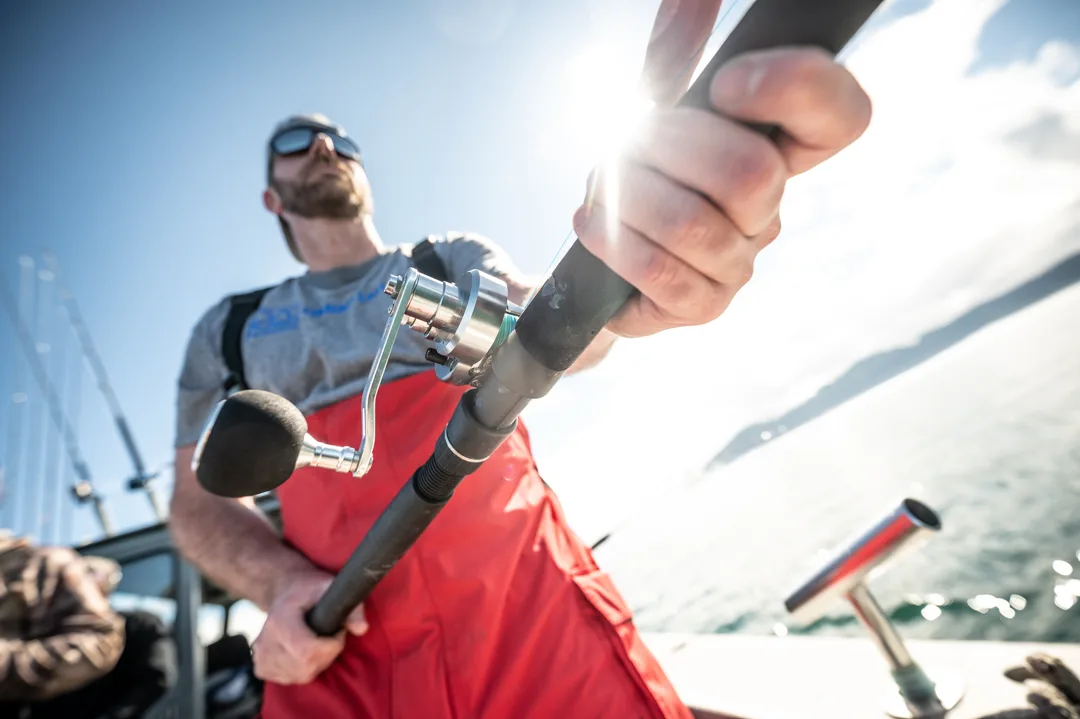 Angler in red bib overalls grips a heavy spinning rod on deck, sun flaring behind the reel, open water in the background.