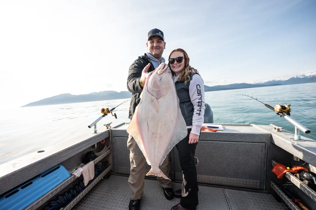 Two anglers holding a large halibut on the deck of a fishing boat, calm water and coastal mountains behind.