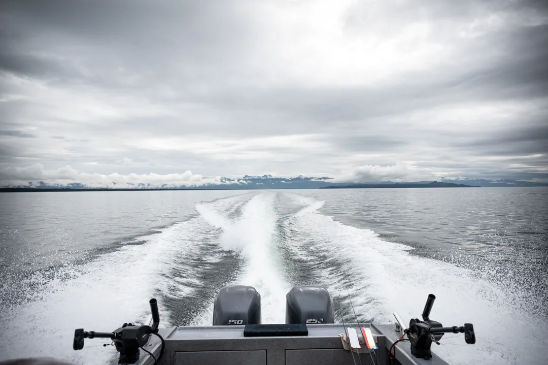 Twin outboards trailing a white wake across Icy Strait, snow-capped mountains on the horizon under overcast skies.