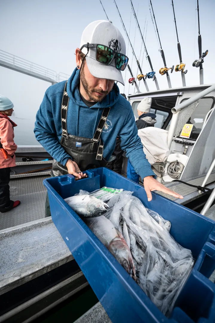 Guide in Helly Hansen bib overalls organizes packaged fish in a blue tote on the boat deck, rods racked behind him.