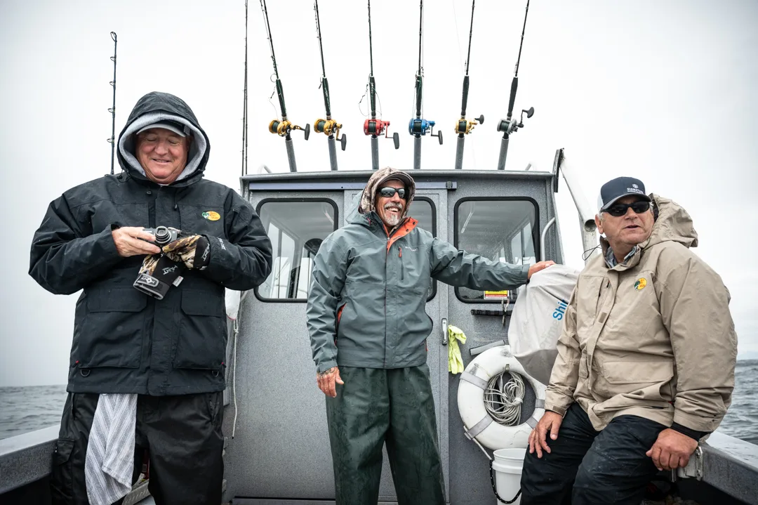 Three anglers in rain gear on the stern deck, rod rack and aluminum wheelhouse behind them, overcast open water.