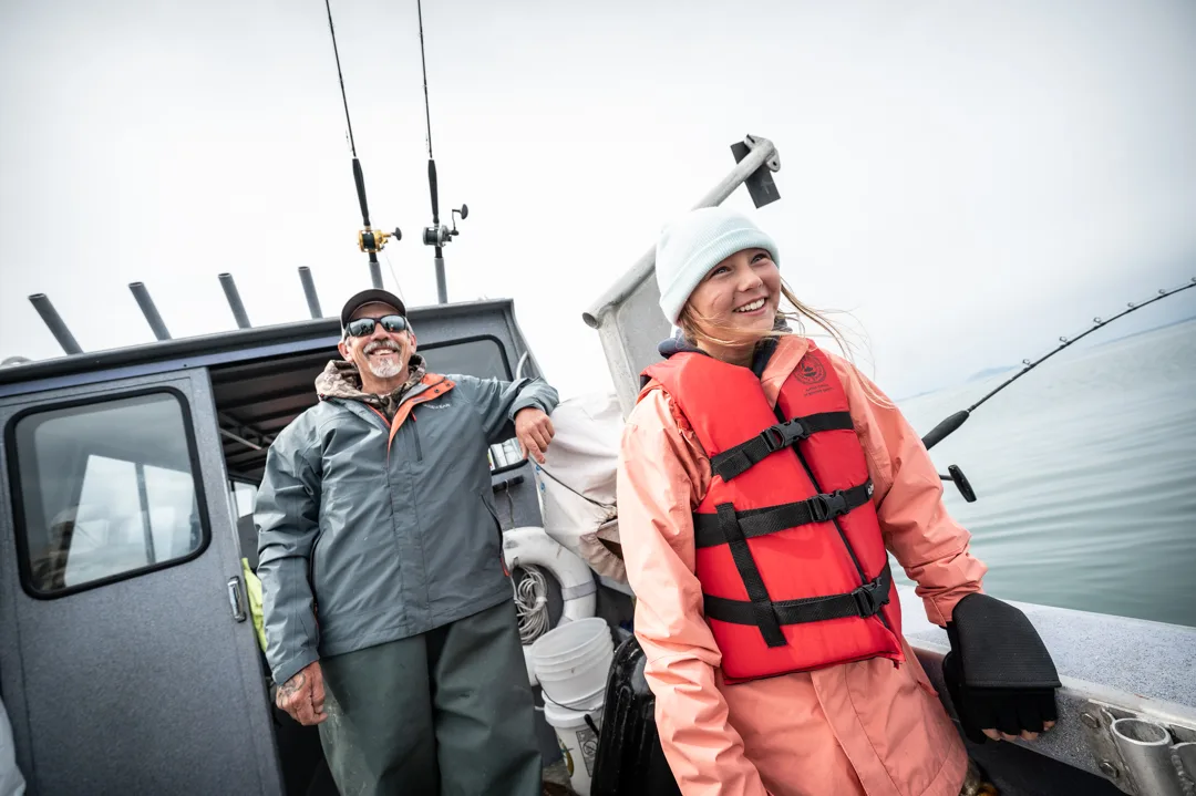 Young woman in red life vest and man in rain gear laughing on the deck of an aluminum fishing boat, rods rigged behind them.