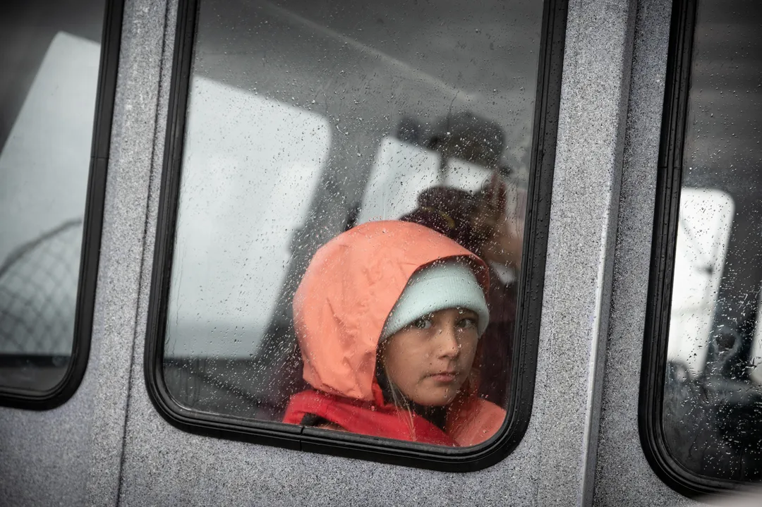 Child in orange rain gear and knit hat looking out a rain-speckled wheelhouse window on an overcast day.