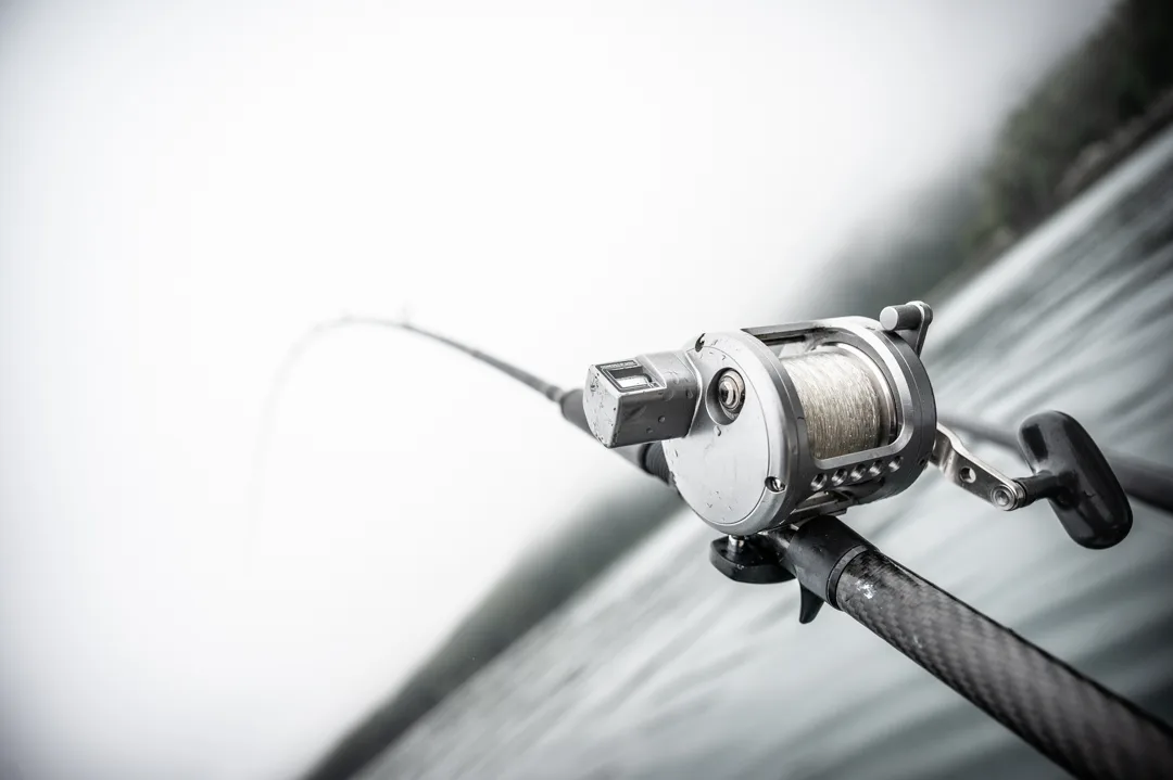 Close-up of a conventional fishing reel and rod in a rod holder, overcast water and shoreline blurred behind.