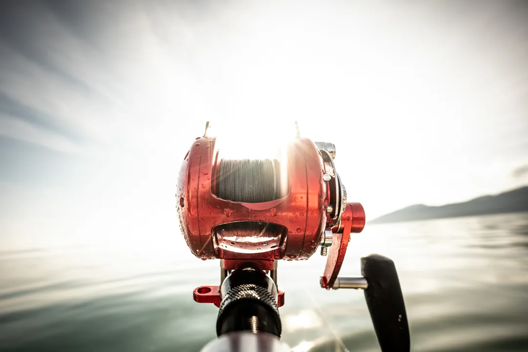 Red baitcaster reel loaded with braided line, water droplets on the spool, calm strait and coastal mountains behind.