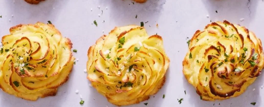 Three golden duchess potatoes piped in rosette form, garnished with chopped herbs and flaked salt on a white surface.
