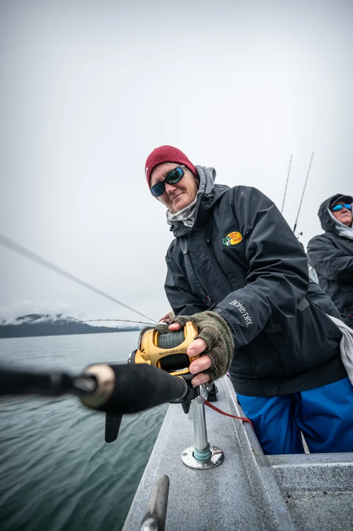 Angler in rain gear works a heavy reel on the boat rail, snow-capped mountains visible across the water behind her.