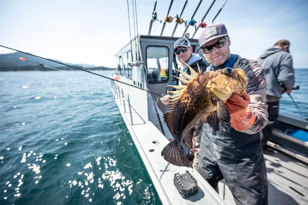 Angler holds a spiny rockfish toward camera on the deck of a charter boat, two other anglers fishing in background.