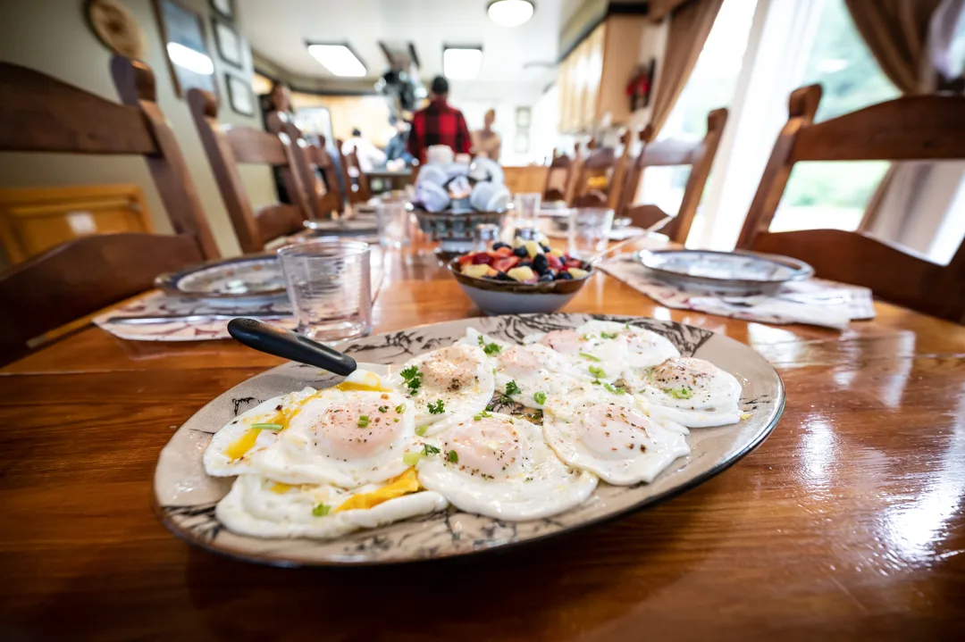 Platter of fried eggs garnished with herbs on the lodge dining table, fruit bowl and guests visible in background.
