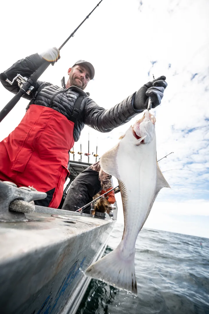 Angler in red rain gear lifts a halibut over the gunwale by the hook, rod bent, open water behind.