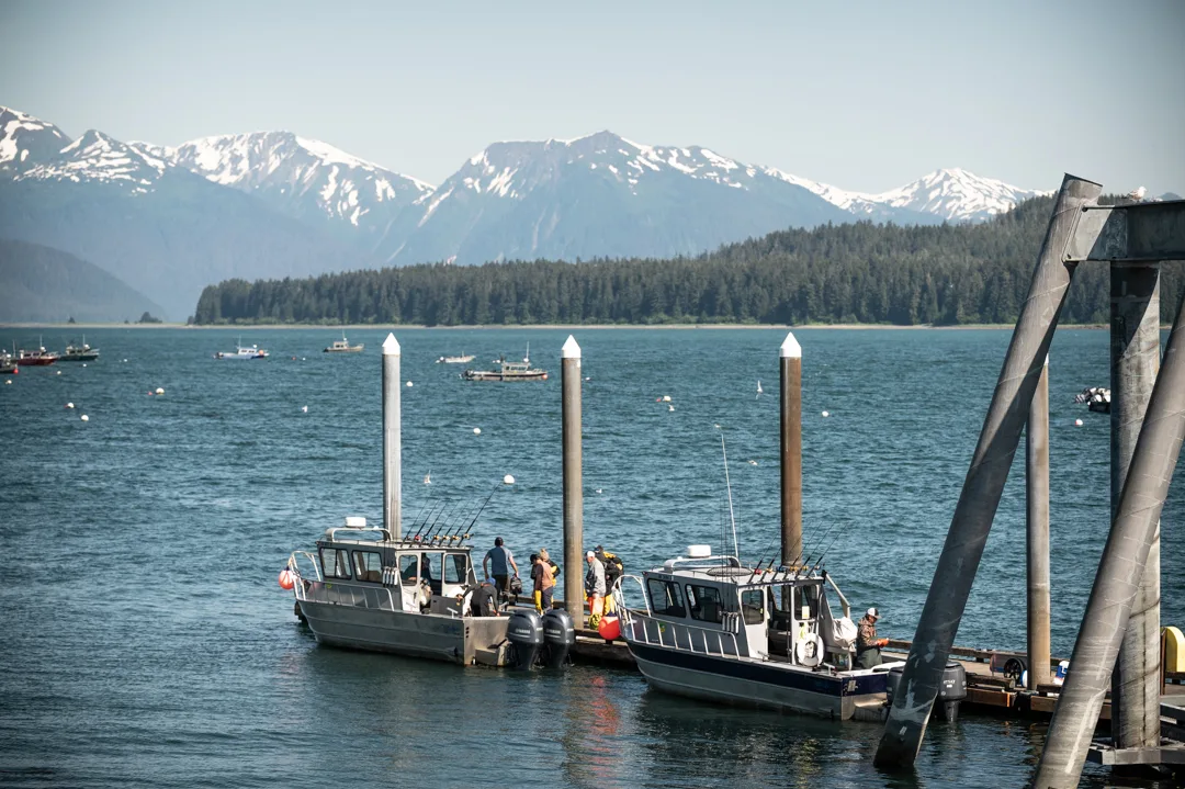 Two aluminum charter boats at a dock in Icy Strait, anglers loading gear, snow-capped mountains across the water.