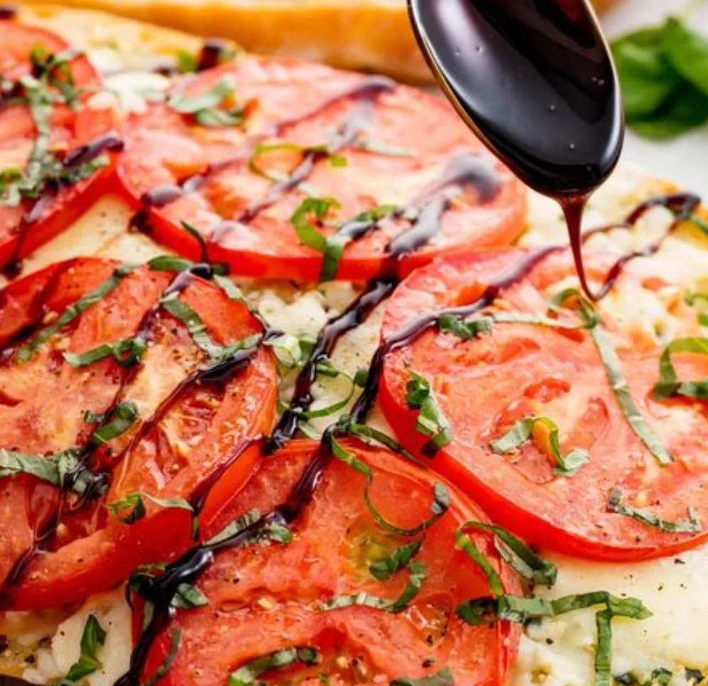 Balsamic glaze drizzled from a spoon over flatbread topped with sliced tomatoes, mozzarella, and fresh basil.