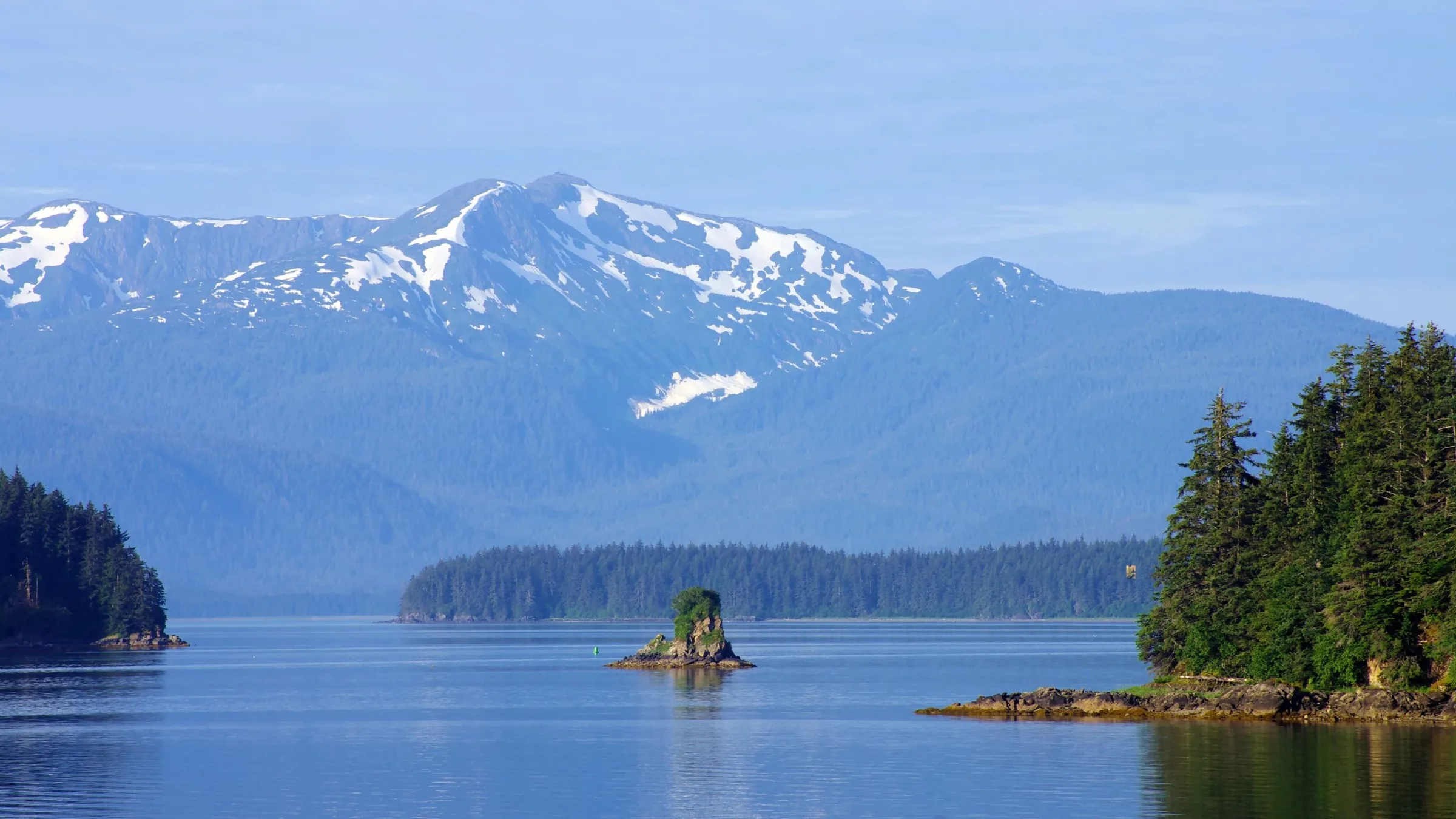 Snow-capped coastal mountains rise above calm blue water near Gustavus, with a small rocky islet and spruce-lined shores in the foreground.