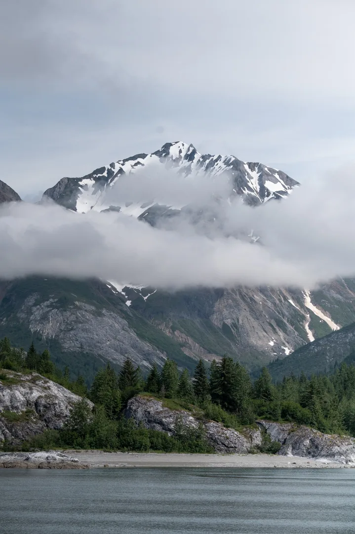 Snow-capped peak rising above low clouds over a rocky shoreline and spruce treeline, Glacier Bay area