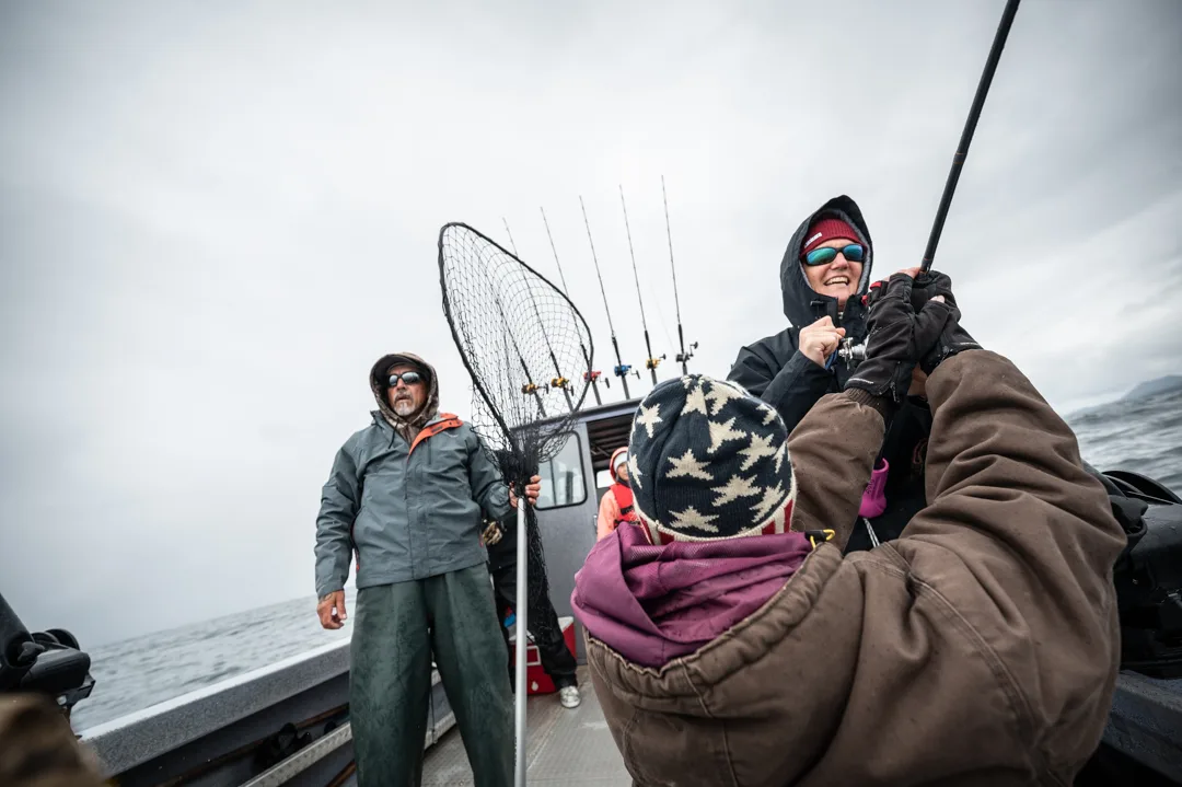 Angler fights a fish on deck while guide stands ready with landing net, overcast Icy Strait in background.