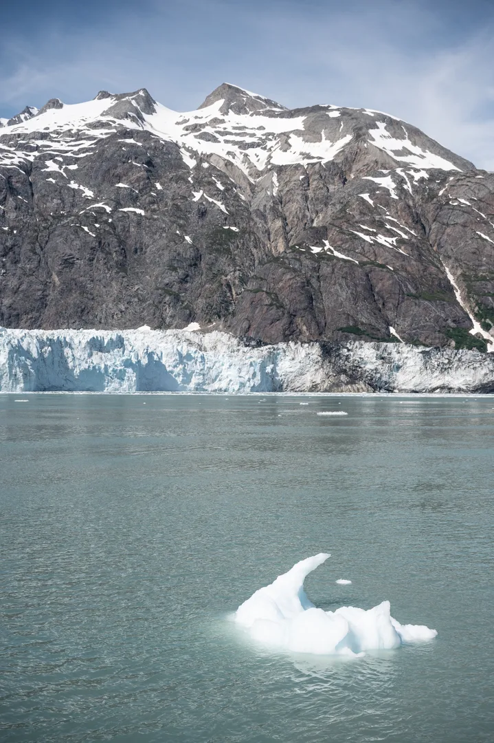 Small iceberg floating in front of a tidewater glacier face, snow-capped mountains rising behind, Glacier Bay, Alaska.