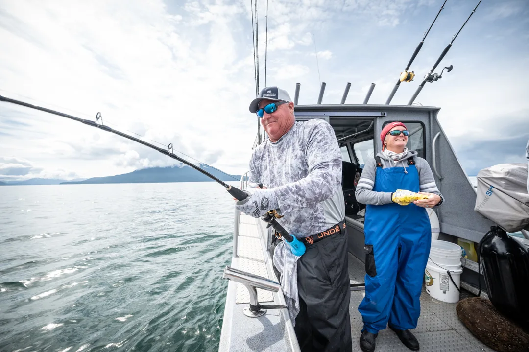 Angler working a bent rod on the stern of a charter boat, guide in blue bibs looking on, mountains across the water.