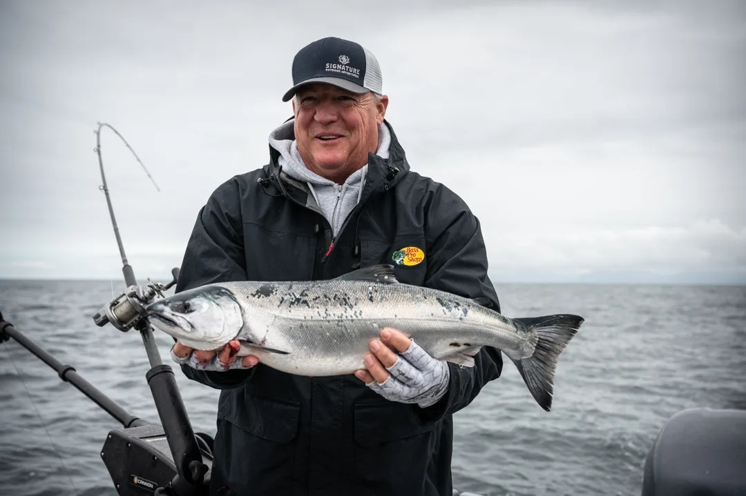 Angler holds a silver salmon on an overcast day on the water, rod and reel visible in background.