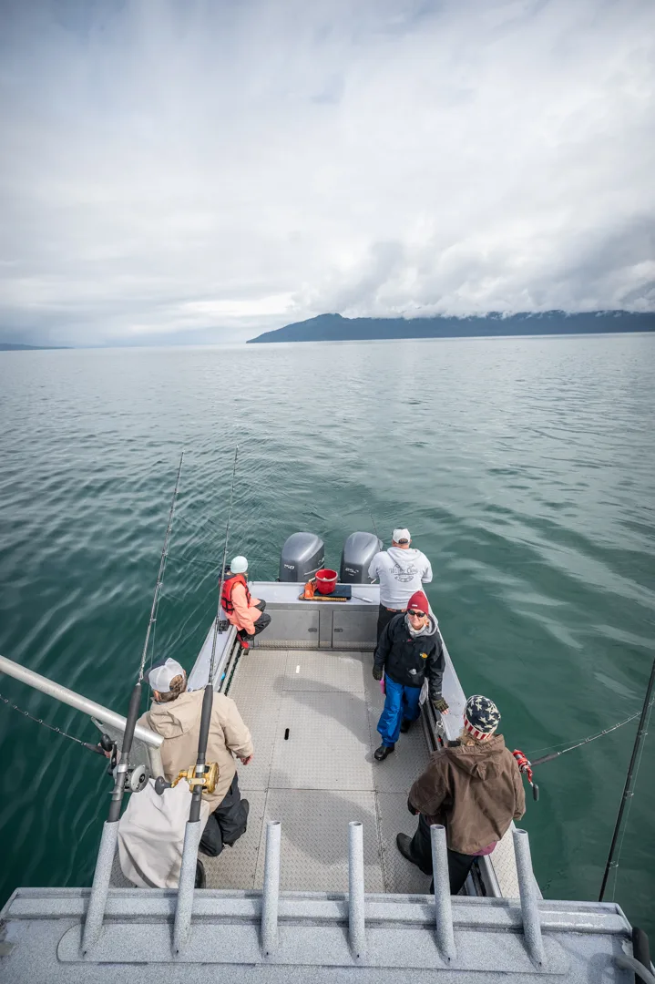 Anglers on the stern deck of an aluminum fishing boat, rods deployed, cloud-draped coastal mountains in the distance.