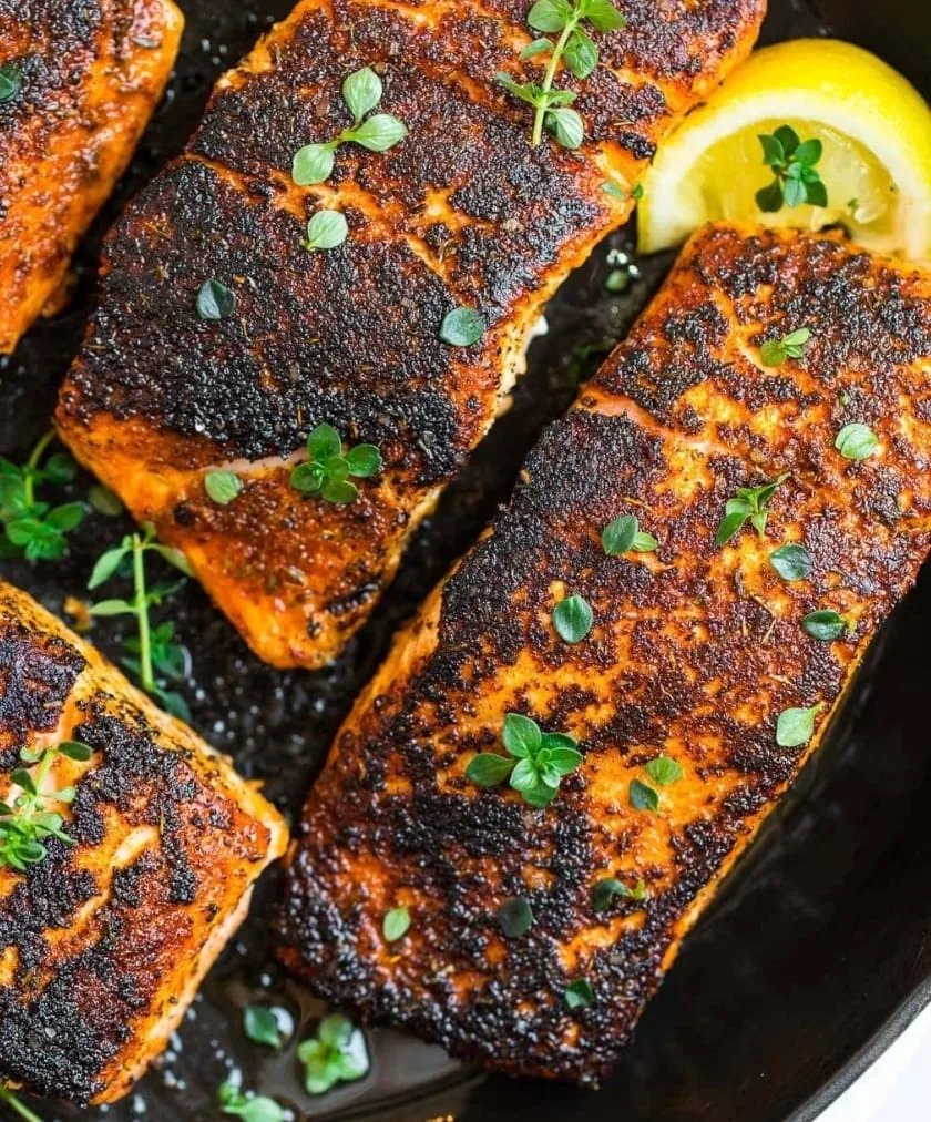 Blackened salmon fillets in a cast iron skillet, scattered with fresh oregano and a lemon wedge.
