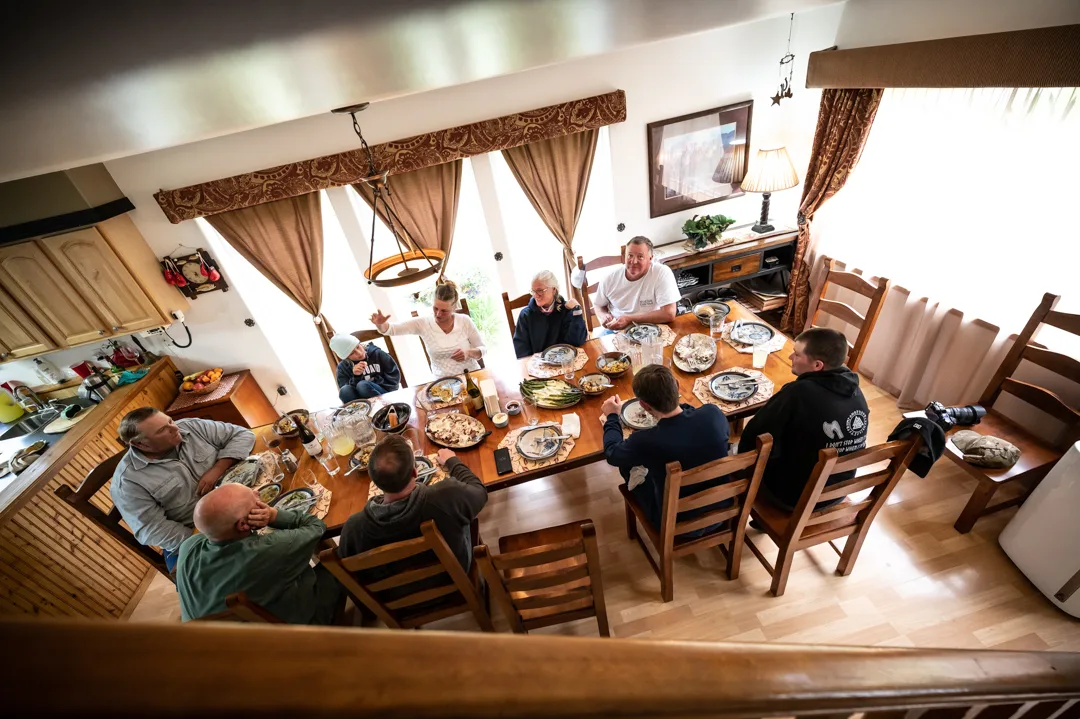 Group of guests gathered around a large dining table for a meal inside the lodge, shot from overhead loft angle.