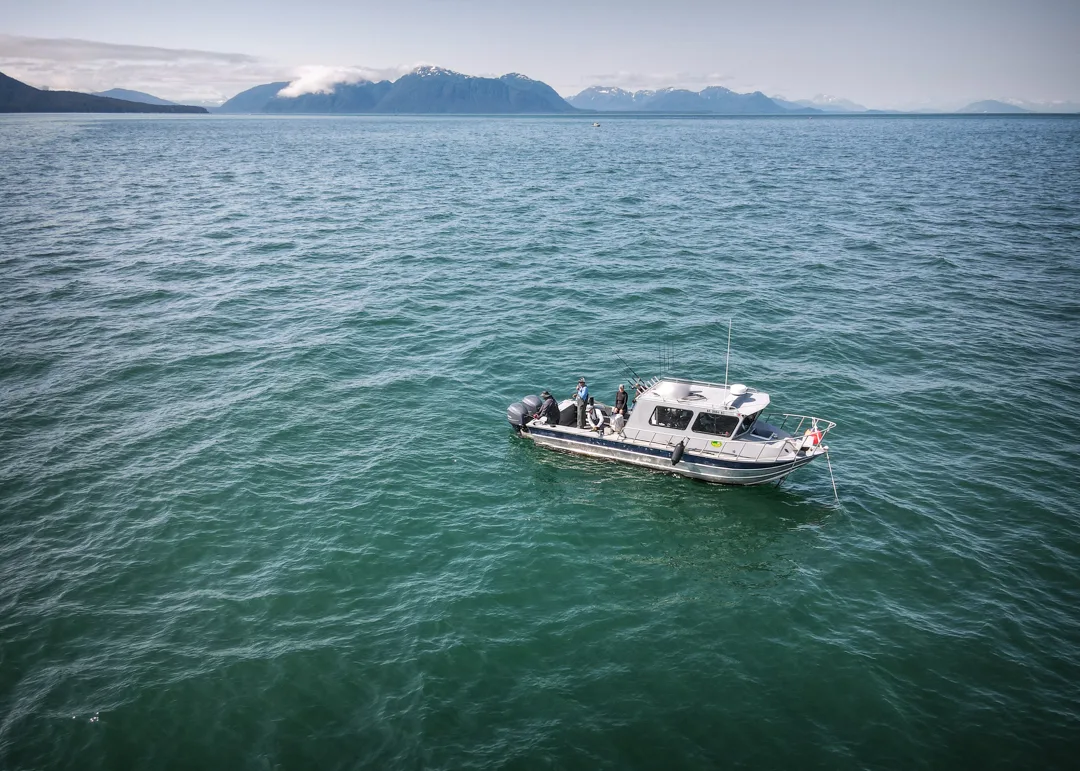 Aerial view of an aluminum charter boat with anglers fishing open water, snow-capped mountains and forested ridgeline in the distance.
