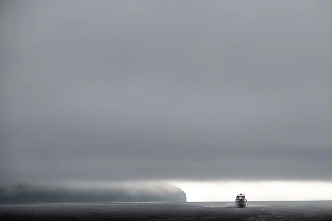 Small fishing boat crossing flat gray water beneath a low fog bank, forested headland dissolving into mist.