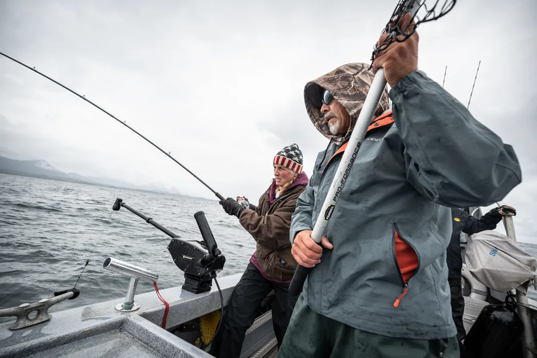Two anglers working rods on an aluminum boat deck in overcast conditions, mountains visible in the mist behind them.