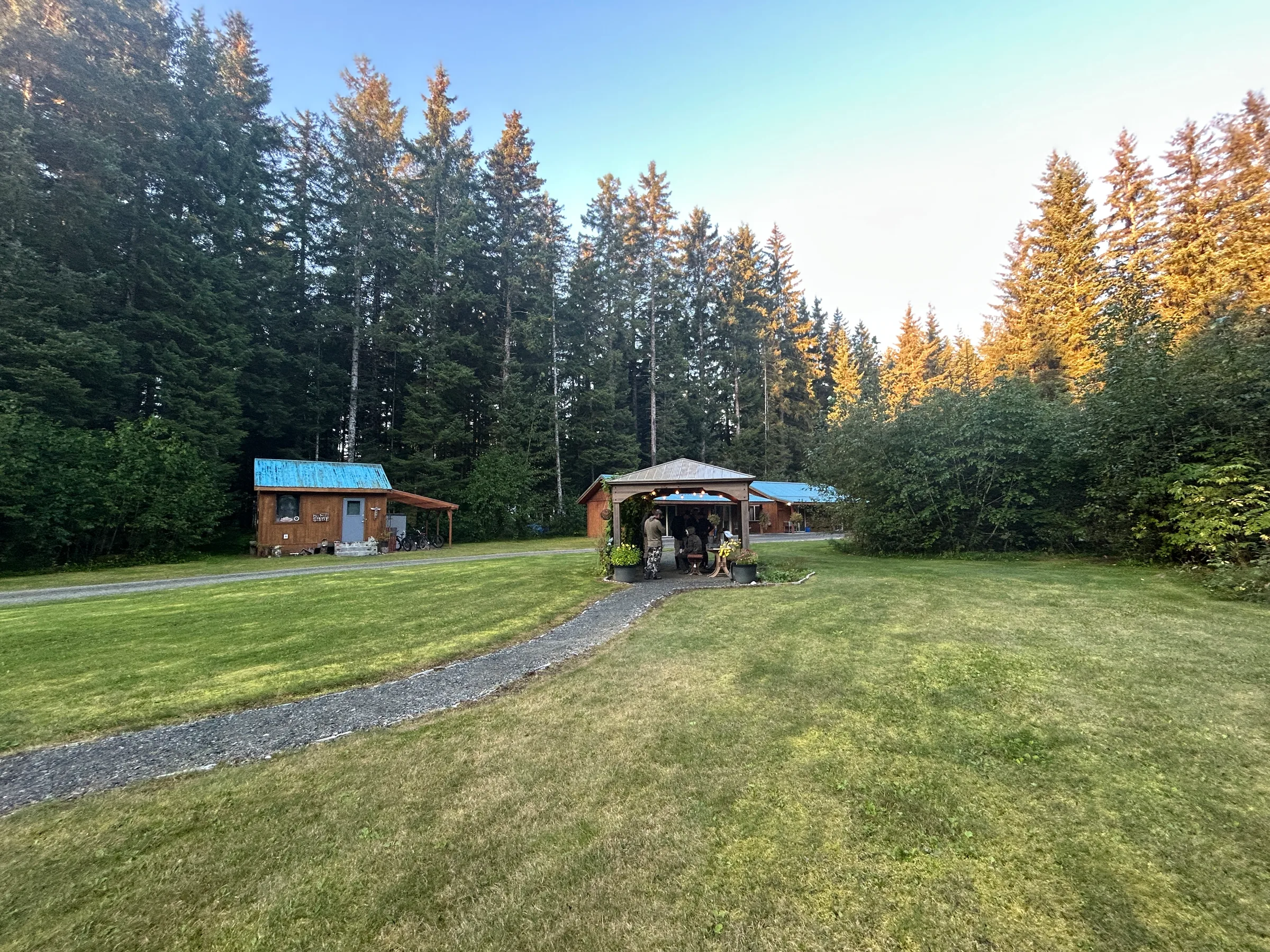 Lodge entry structure and outbuilding on a gravel path, surrounded by spruce forest in warm evening light, Gustavus Alaska.
