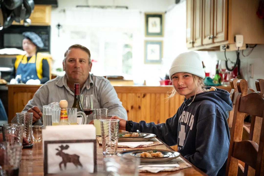 Man and young girl seated at a wood dining table inside the lodge, chef visible working in the kitchen background.