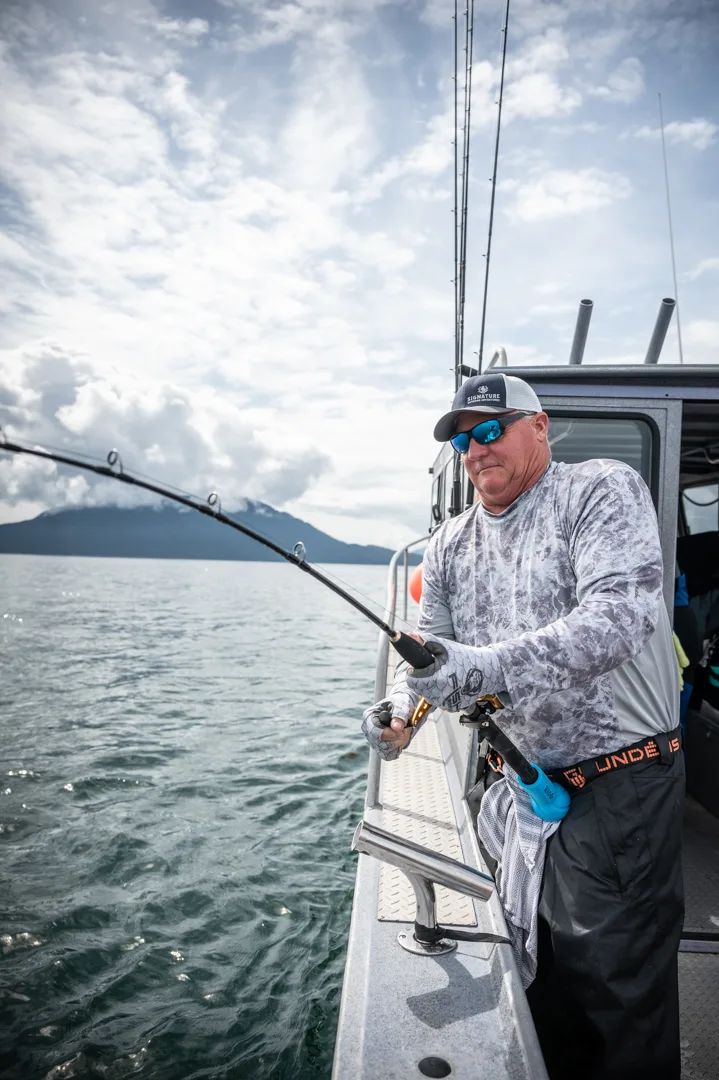 Angler working a bent rod off the stern rail, mountains and open water visible behind him.