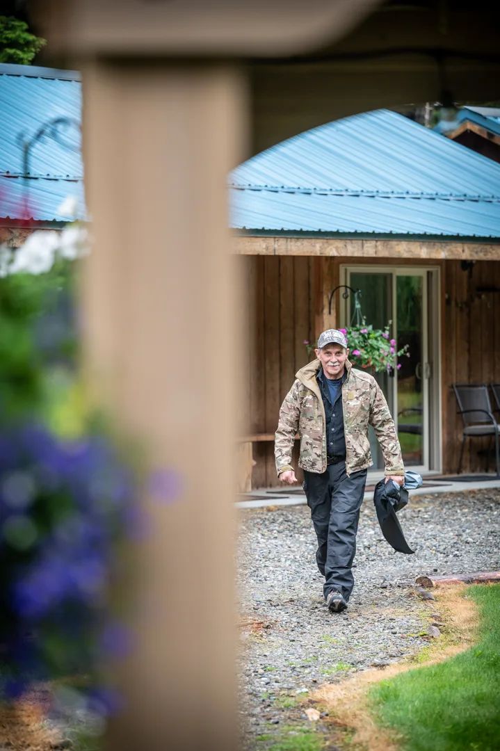 Guest in camo jacket walking gravel path toward lodge cabin with blue metal roof and hanging flower baskets.